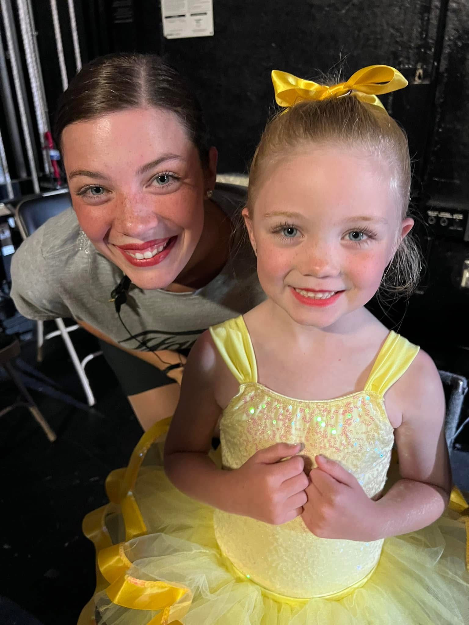 A teenage dancer crouching down beside a small dancer in a yellow tutu and matching hair bow, backstage