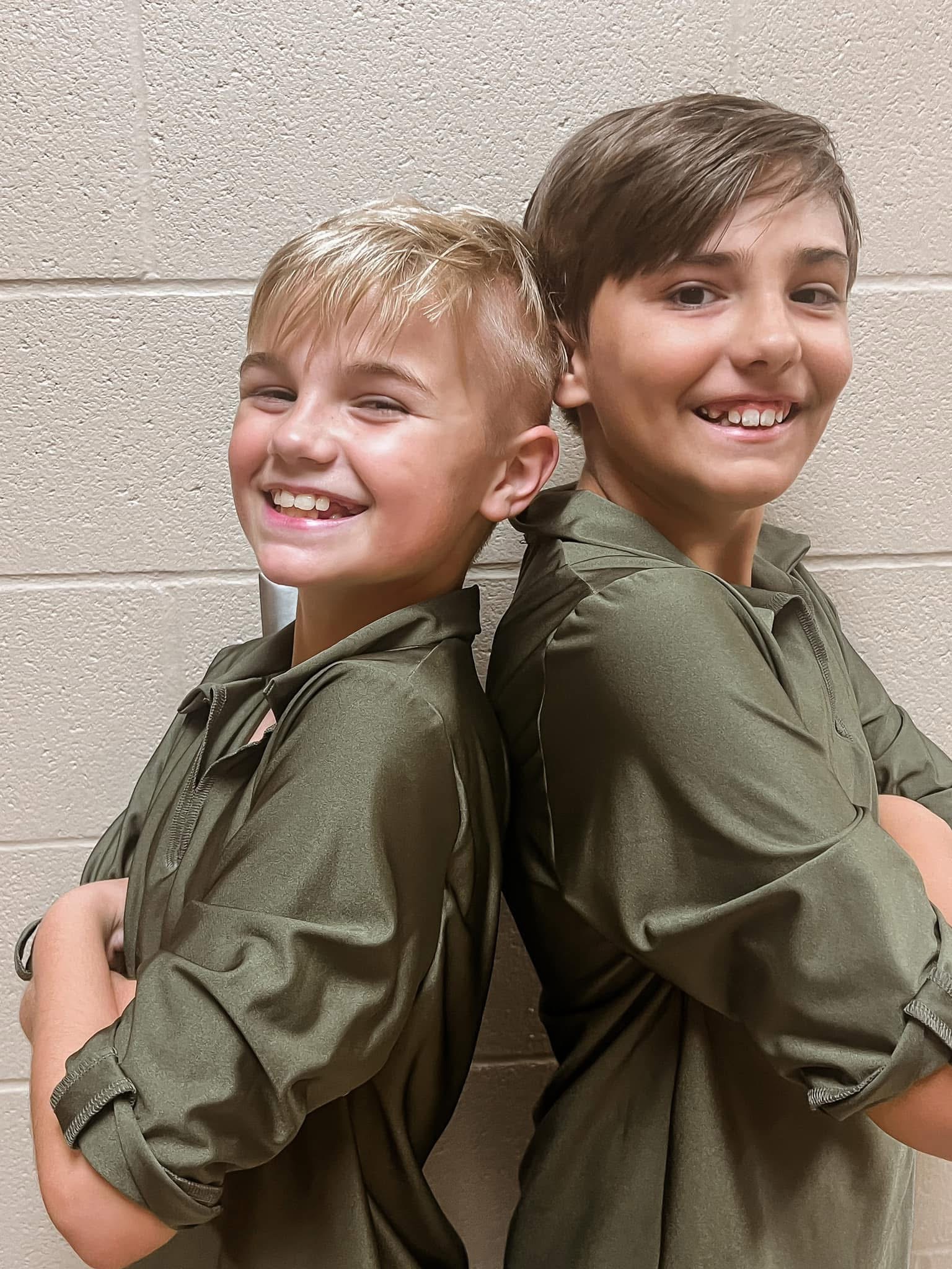 Two young dancers standing back to back in matching olive costumes, laughing
