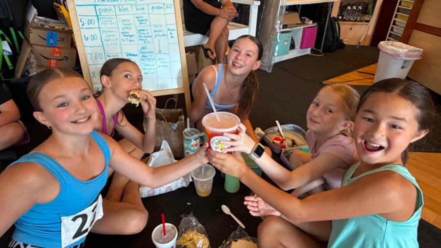 Dancers sitting on the studio floor sharing food between classes, the schedule board behind them