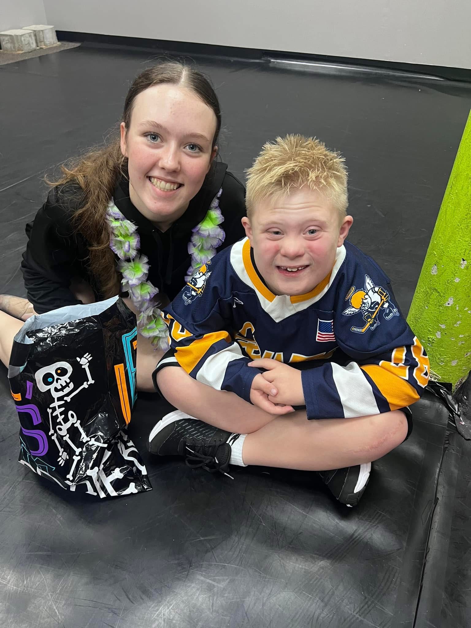 A teenage dancer sitting on the studio floor beside a younger dancer in a hockey jersey, both grinning at the camera