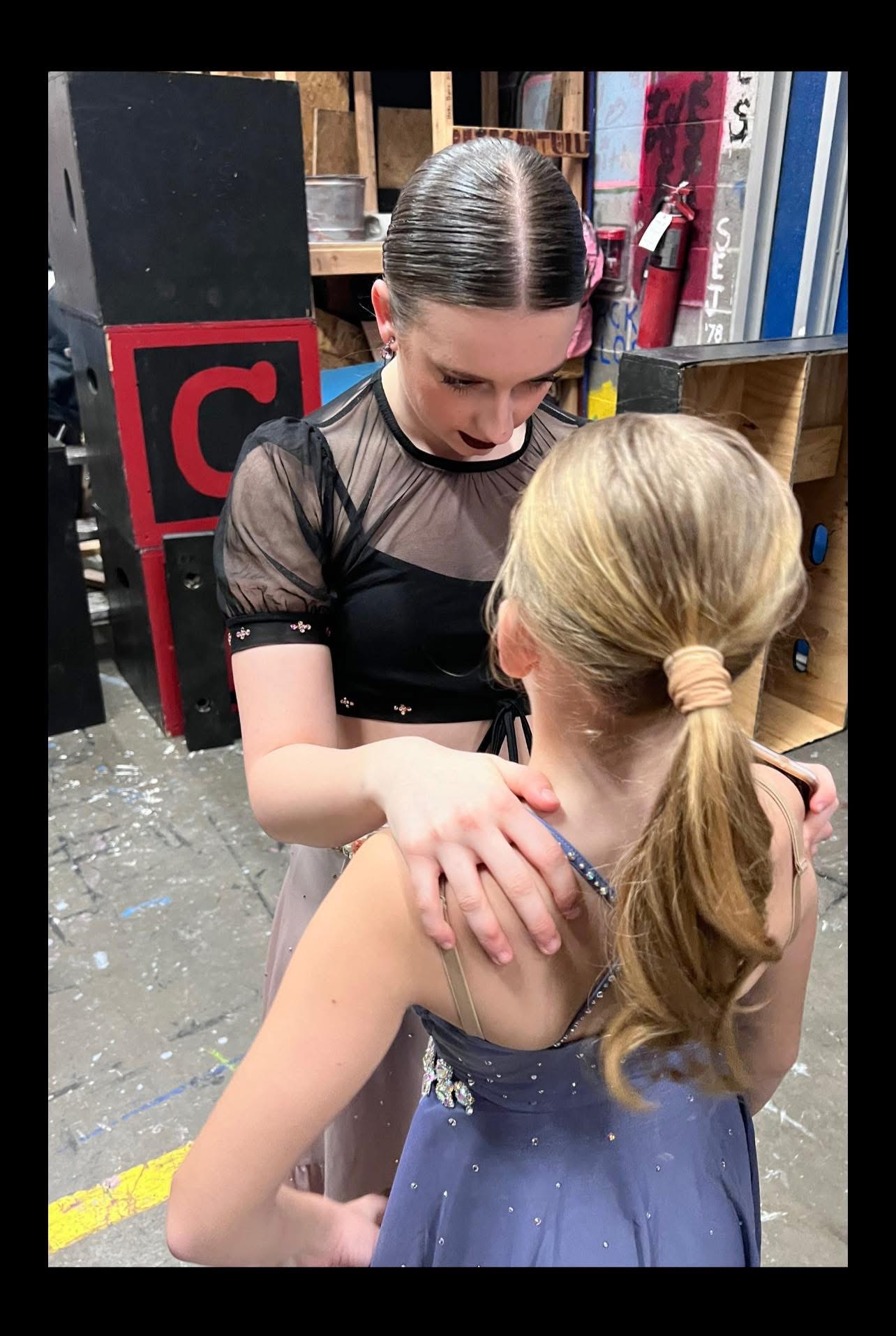 An older dancer fastening a younger dancer’s costume backstage before they go on