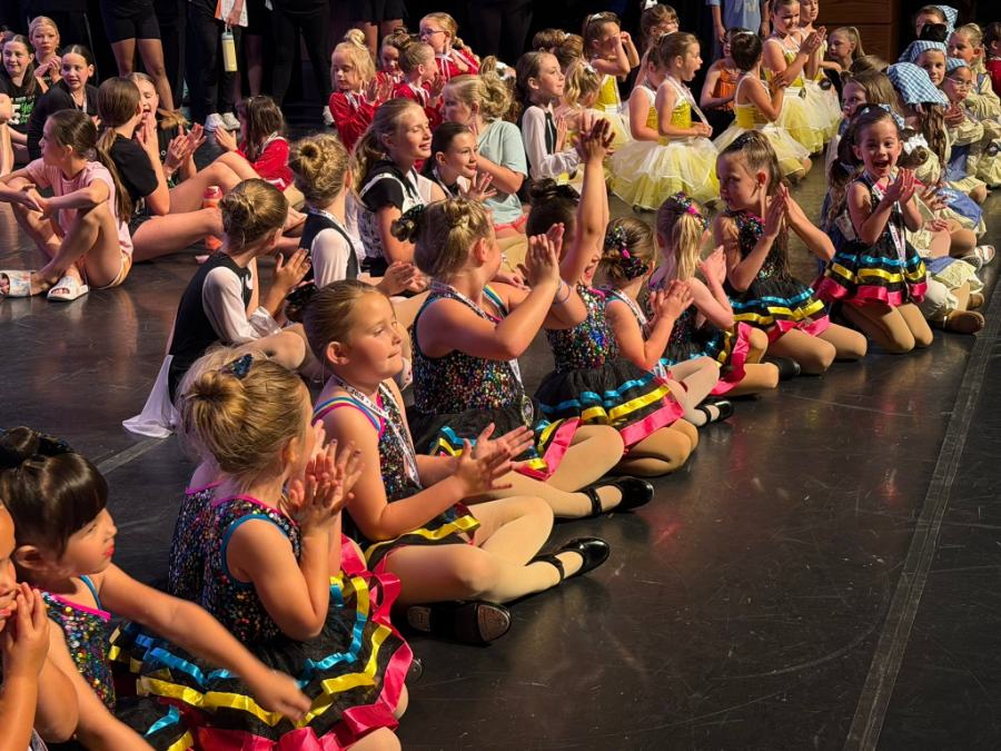 Rows of young dancers in costume sitting on the recital stage, clapping