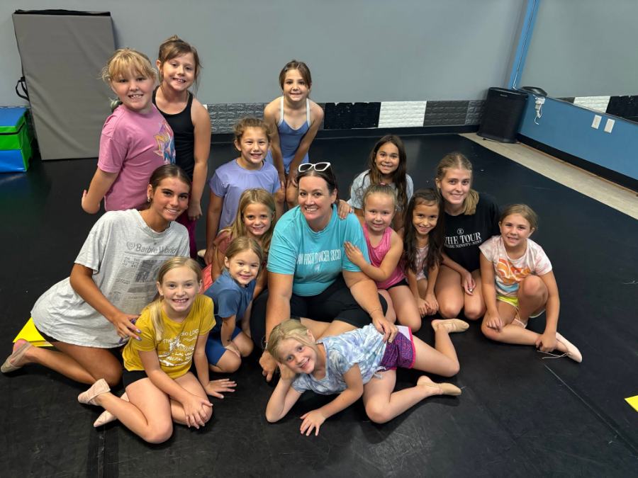 A teacher sitting on the studio floor surrounded by her whole class of young dancers, everyone laughing