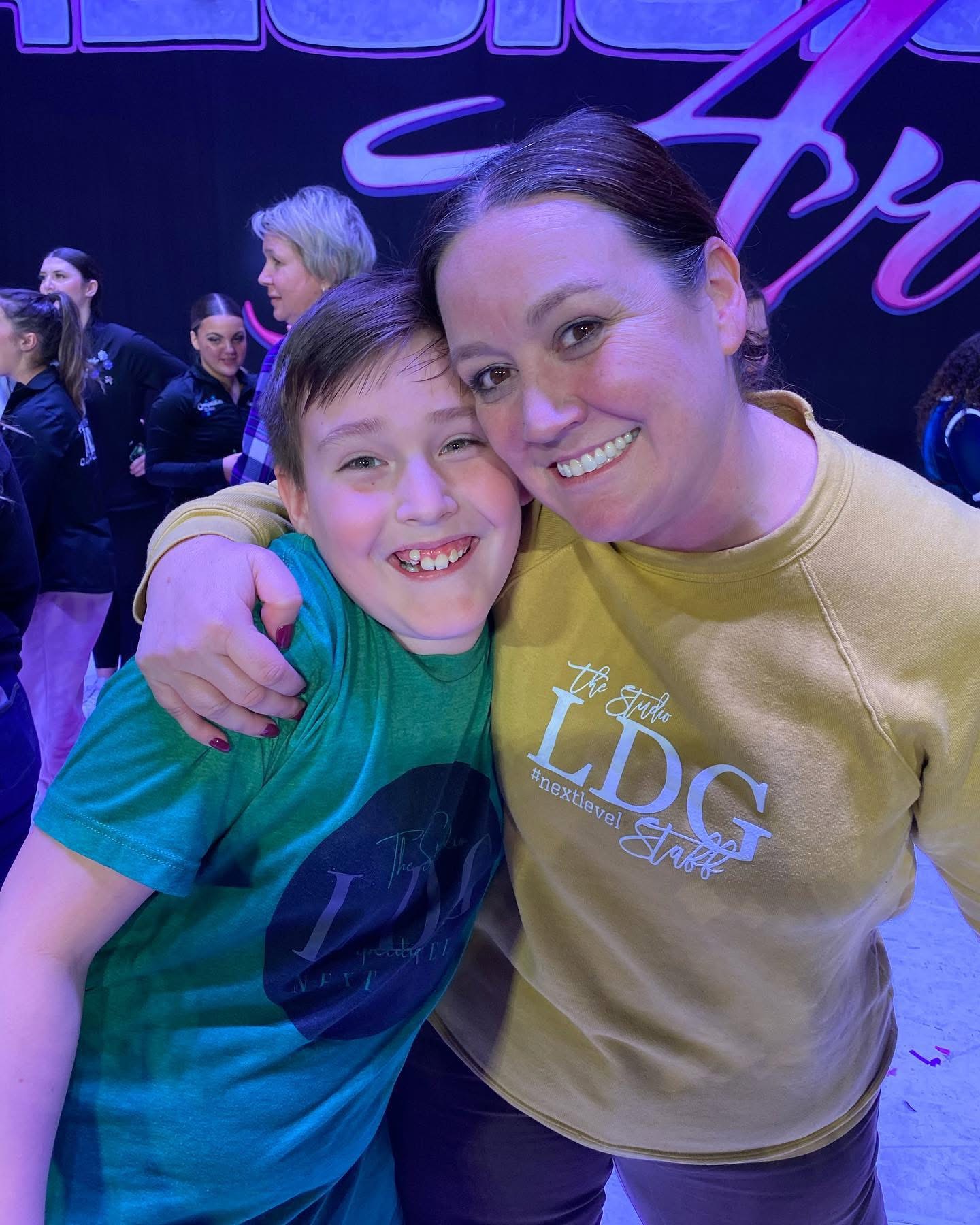A young dancer and a teacher in an LDG staff sweatshirt, cheek to cheek at a competition