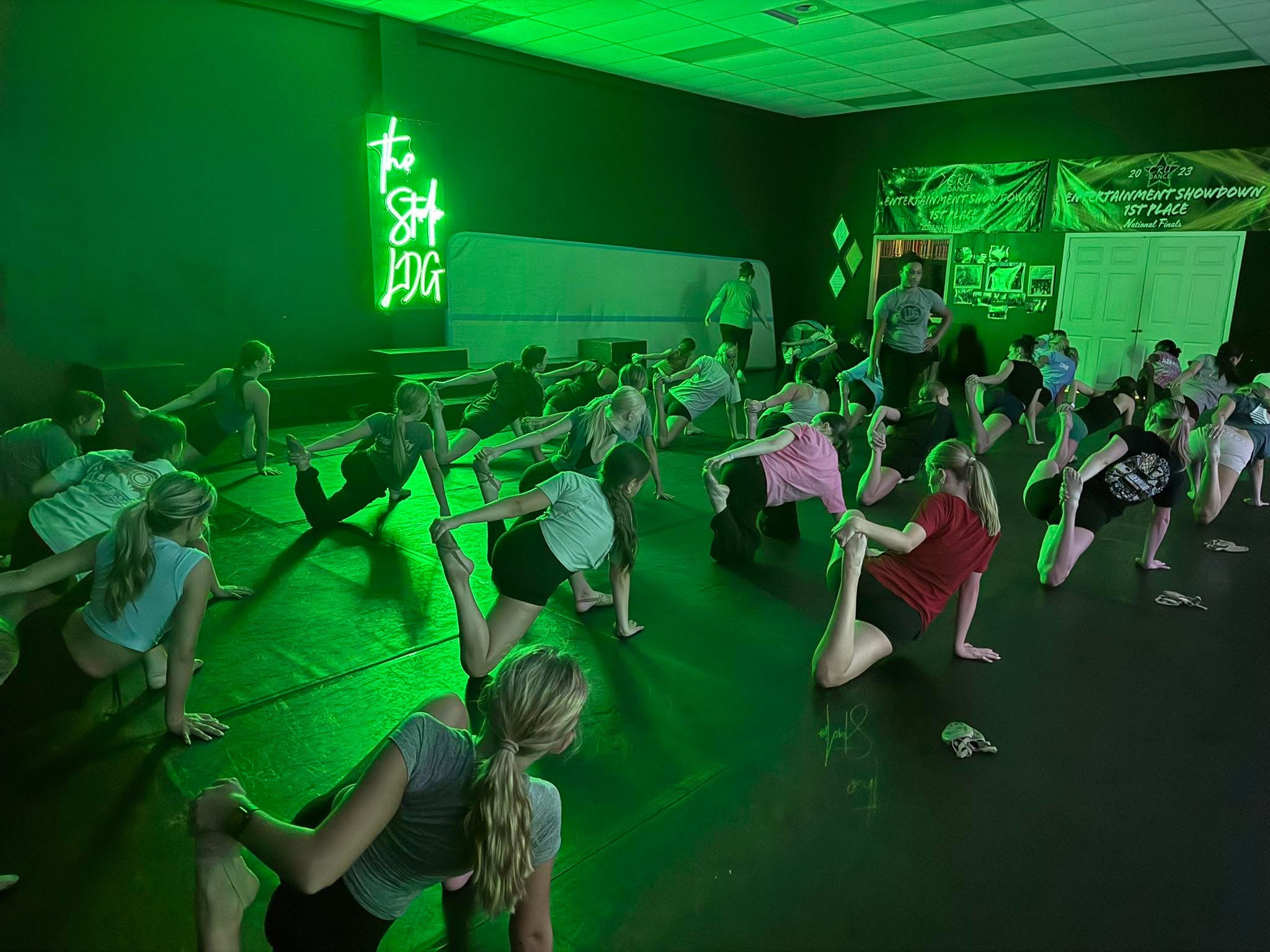 A whole class stretching on the studio floor under green light, with the neon The Studio LDG sign lit on the wall behind them